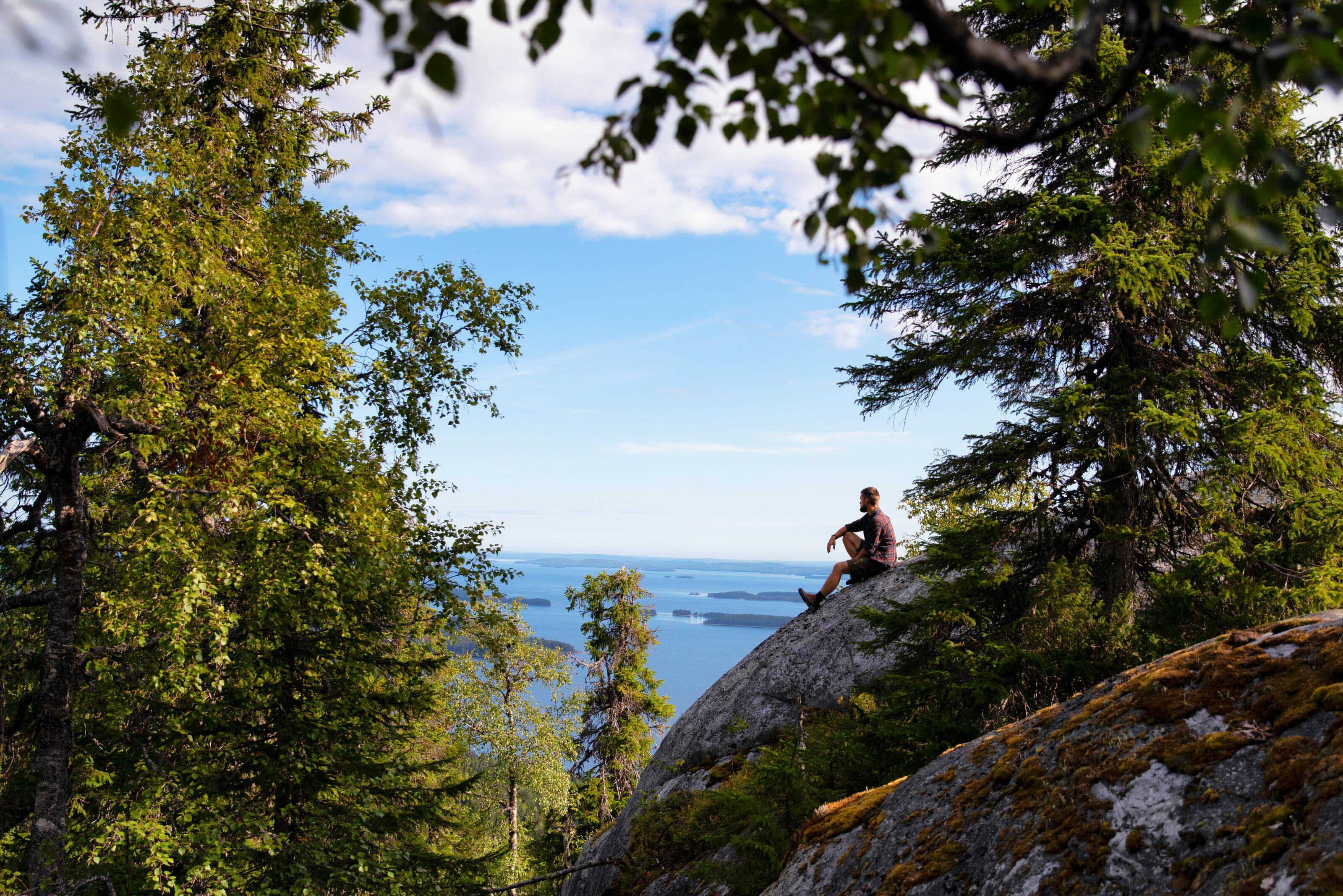 Ein Mann sitzt auf einem Felsen im Koli-Nationalpark und zu seinen Füßen breitet sich die finnische Seenlandschaft aus